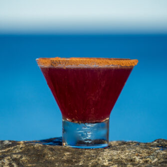 Pomegranate Cosmopolitan, a refined cocktail with Earl Giles Vodka, Tattersall Orange Crema, Fresh-Squeezed Lime, and Pomegranate Molasses, is enjoyed against a serene waterfront backdrop.